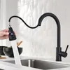A black kitchen faucet with a pull-down sprayer is shown in use over a stainless steel sink. A hand is holding the sprayer, and water is flowing out. Produktbild-Vorschau