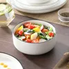 A close-up of a salad in a white bowl with a black rim, set on a wooden table. Other tableware and a glass of water are visible in the background. Náhled obrázku produktu