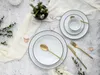 A flat lay of a white porcelain dinnerware set with black trim, accented with gold cutlery and a small floral arrangement. Podgląd zdjęcia produktu