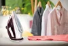 An iron is placed on an ironing board next to a piece of clothing with a zipper. In the background, several shirts are hanging on hangers. Produktbild-Vorschau