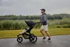A man is jogging with a baby stroller on a paved path next to a grassy area and a body of water. Náhled obrázku produktu