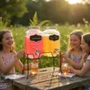 Three young girls are sitting at a wooden table outdoors, with two beverage dispensers filled with pink and yellow drinks. The sun is setting in the background. Náhľad obrázku produktu