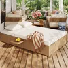 A rattan garden sofa set with white cushions and a striped throw blanket on a wooden deck, surrounded by lush greenery and flowers. Anteprima dell’immagine prodotto