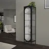 A tall, narrow black display cabinet with glass doors and shelves, filled with white dishes, stands in a modern living room next to a white armchair. Podgląd zdjęcia produktu