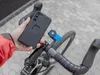 A person is holding a black smartphone case with a Quad Lock mount attached, about to attach it to a bicycle handlebar mount. Náhled obrázku produktu