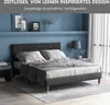 A dark gray upholstered bed with light gray bedding is shown in a bedroom setting with a blue wall and white curtains. Produktbild-Vorschau