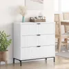 A white dresser with three drawers is shown in a room with a plant and a dining table in the background. Náhled obrázku produktu