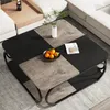 A square coffee table with a two-tier design, featuring a mix of black and wood-grain finishes, placed in a living room setting with a sofa in the background. A small bowl of oranges and a book are on the table. Podgląd zdjęcia produktu