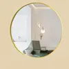A round gold-framed mirror reflecting a modern living room with a green chair and pendant lights. Produktbild-Vorschau
