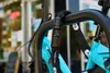 A close-up shot of a black combination chain lock securing a teal bicycle to a black bike rack. The lock has a digital keypad and is wrapped around the bike frame and the rack. The ABUS logo is visible on the chain cover. Produktbild-Vorschau