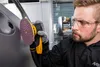 A man wearing safety glasses and gloves is using a sander on a car door. Produktbild-Vorschau