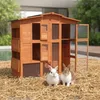 A wooden rabbit hutch with two rabbits in front of it, set on a bed of straw, with a pool and trees in the background. Náhled obrázku produktu