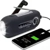 A black emergency radio with a flashlight and antenna is shown next to a smartphone that is charging. Produktbild-Vorschau