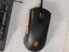 A black gaming mouse with orange accents is placed on a white grid surface next to a portion of a keyboard. Náhled obrázku produktu