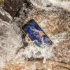 A rugged smartphone is submerged in flowing water, surrounded by rocks. Produktbild-Vorschau