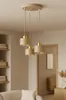 A modern beige pendant light fixture with three lamps hangs in a well-lit room, possibly a living room or dining area, with shelves and furniture visible in the background. Produktbild-Vorschau