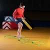 A woman is performing agility drills on a yellow mat, stepping through hexagonal agility rings. In the background, there are stacked agility boxes with colorful designs. Náhled obrázku produktu