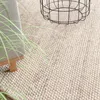A close-up view of a round, textured rug with a light beige and white speckled pattern. A black wire basket and a portion of a grey upholstered chair with wooden legs are visible on the rug, along with some green grass-like foliage. Produktbild-Vorschau