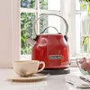 A red KitchenAid electric kettle sits on a countertop next to a cup and saucer. A bowl of strawberries and a vase with flowers are visible in the background. Produktbild-Vorschau