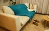 A cozy living room scene with a blanket draped over a sofa, slippers on the floor, and a small coffee table. Produktbild-Vorschau