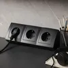 A black power strip with three outlets is shown on a desk, with a plug inserted into one outlet. Headphones and a notebook are also visible. Náhled obrázku produktu