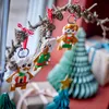Christmas-themed LEGO gingerbread figures hanging from a branch with pinecones, with decorative Christmas trees and gift boxes in the background. Produktbild-Vorschau