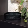A black washbasin with a ribbed texture sits on a black countertop, with a gold faucet behind it. A large green leaf is to the right of the faucet. Produktbild-Vorschau