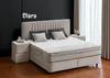 A bedroom scene featuring a beige box spring bed with two pillows and a headboard, set against a black slatted wall and next to a beige nightstand. Curtains are visible in the background. Produktbild-Vorschau