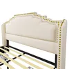 A close-up view of a beige upholstered bed frame with gold nailhead trim. Produktbild-Vorschau