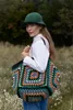 A woman wearing a green bucket hat and a white shirt stands in a field, holding a large, colorful crocheted tote bag. Produktbild-Vorschau
