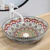 A decorative basin with a colorful mandala pattern, placed on a wooden surface with a white faucet. A mirror is visible in the background. Náhled obrázku produktu
