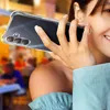 A woman is holding a clear phone case with a Samsung Galaxy A16 phone inside, up to her ear as if on a call, in a blurred outdoor setting. Produktbild-Vorschau