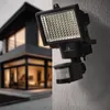 A black LED floodlight with a motion sensor is mounted on a textured wall, with a modern house blurred in the background. Produktbild-Vorschau