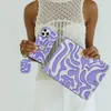 A person is holding a phone case, an AirPod case, and a laptop sleeve, all with a matching purple and white wavy pattern. Podgląd zdjęcia produktu