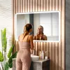 A woman is applying makeup in front of a large, illuminated mirror in a modern bathroom. Produktbild-Vorschau