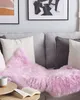 A pink sheepskin rug is draped over a gray sofa with several pillows. A book and a mug are on a wooden side table in the foreground. Produktbild-Vorschau