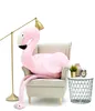 A large pink flamingo plush toy is sitting in a beige armchair next to a small side table with decorative items. Produktbild-Vorschau