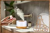 A hand holding a small bottle of essential oil over an aroma diffuser that is emitting mist. The scene is set on a countertop with a plant and another bottle in the background. Produktbild-Vorschau