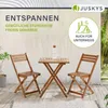 A wooden bistro set with two chairs and a table on a patio, with plants and a wall in the background. Produktbild-Vorschau