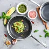 A top-down view of a frying pan with zucchini noodles and pesto, alongside ingredients like garlic, basil, and chili peppers. Náhľad obrázku produktu