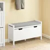 A white bench with a gray cushion is shown in a room with a yellow wall and a rug on the floor. A gray coat is hanging on the wall above the bench. Produktbild-Vorschau