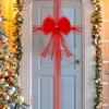 Große rote Tüllschleife mit funkelnder Mitte, vertikal und horizontal um eine hellblaue Tür gewickelt, flankiert von geschmückten Weihnachtsbäumen und Girlanden Podgląd zdjęcia produktu