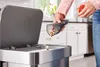 A person is emptying a handheld vacuum cleaner into a trash can in a kitchen. Produktbild-Vorschau