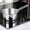 A stainless steel saucepan with a long handle sits on a reflective black surface, possibly a stovetop. The background is blurred, showing hints of greenery and jars. Produktbild-Vorschau