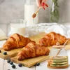 A hand is drizzling honey from a dipper onto croissants, with a glass of milk and a honey jar in the background. Produktbild-Vorschau