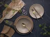 A table setting with plates, cutlery, a bowl, and a napkin, arranged on a dark stone surface, with a wooden board and beans in the background, and some greenery. Náhľad obrázku produktu