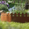 A garden bed border made of rusted metal with a picket fence design, separating a grassy lawn from a flower bed with purple and blue flowers. Produktbild-Vorschau
