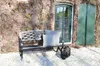 A black garden bench with two patterned cushions sits on a gravel path in front of a brick wall covered in vines. A green door and shutter are visible. Anteprima dell’immagine prodotto