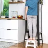 A person is standing on a white step stool in a kitchen, reaching into a glass cabinet. Produktbild-Vorschau