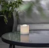 A white LED candle sits on a glass-topped table, with a blurred plant in the background. Produktbild-Vorschau